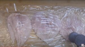 Pounding chicken breasts under plastic wrap with a kitchen mallet on a wooden surface.